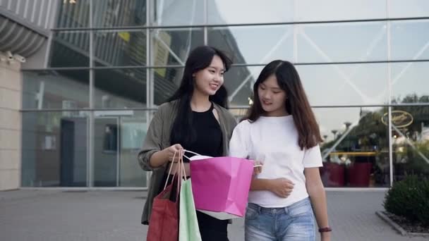 Schöne lächelnde asiatische Frauen, die in der Nähe eines großen Einkaufszentrums spazieren und sich gegenseitig die Einkäufe zeigen, die sie nach dem Einkaufen gekauft haben — Stockvideo