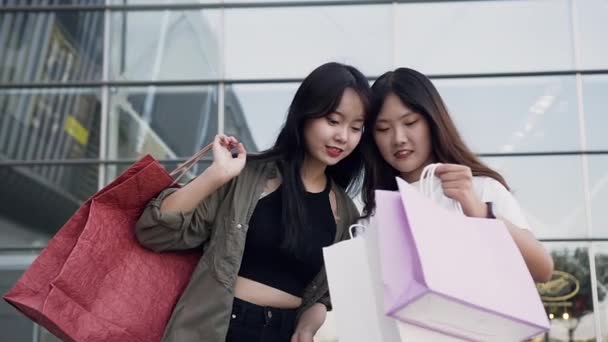 Two joyful likable young asian girlfriends standing near shopping mall and showing each other what they bought after shopping — Stock Video