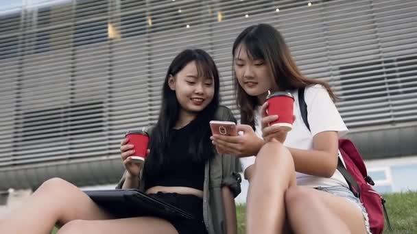 Привлекательные счастливые улыбающиеся азиатские девушки, держащие в руках кофе и смотрящие на телефон, сидящие на траве возле большого городского здания — стоковое видео