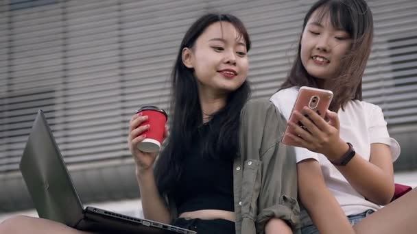 Красиві щасливі усміхнені азіатські дівчата сидять біля великої міської будівлі і використовують телефон для перегляду фотографій — стокове відео