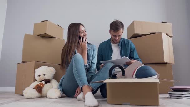 Menina muito feliz falando no telefone enquanto sentado no chão com seu namorado bonito que olhando para papéis com design de seu novo apartamento — Vídeo de Stock