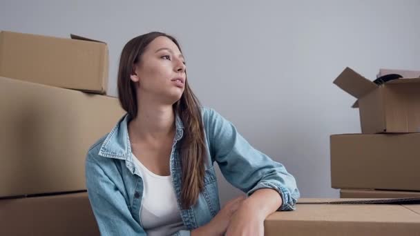 Attractive tired young woman resting on the box in the empty new flat with many boxes after relocation — Stockvideo