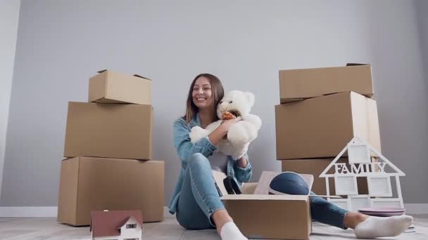 Adorable joven feliz sentada en el suelo y desempacando la caja de cartón después de la reubicación a su nuevo apartamento — Vídeos de Stock