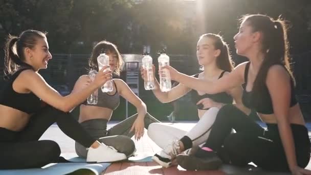 Mooie glimlachende sportieve meisjes in het trainen van kleding zitten op outdoor stadion en geven high five met flessen water — Stockvideo