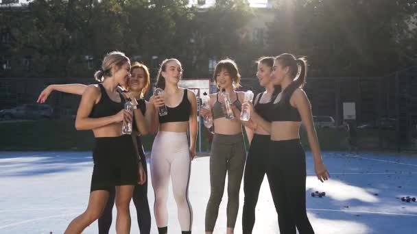 Счастливые улыбающиеся спортивные девушки в тренировочной одежде, стоящие на открытом стадионе и дающие пять бутылок после спортивных тренировок — стоковое видео