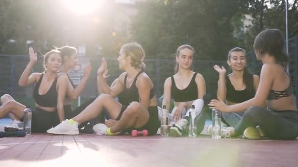 Des femmes souriantes et athlétiques qui célèbrent la fin de l'entraînement de fitness et donnent cinq places assises sur le sol du stade dans le parc — Video