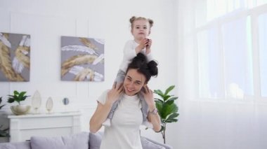 Çekici, mutlu, gülümseyen, komik saç stili olan genç bir kadın. Omzuna sarılmış. Tatmin olmuş küçük kızı, evde kameraya poz veriyor.