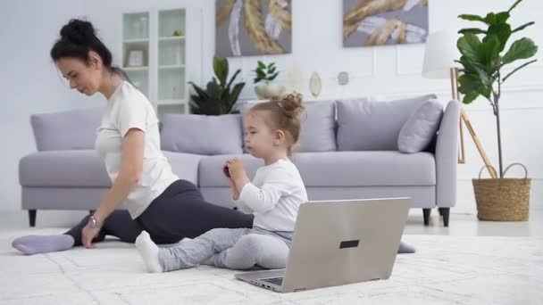Aantrekkelijk klein meisje zitten in de buurt van de laptop op de vloer, terwijl haar zelfverzekerde sportieve donkerharige moeder stretching in de buurt van haar — Stockvideo