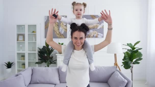 Visão frontal de apto atraente sorrindo jovem mulher ativa que fazendo exercícios de agachamento com sua filha pequena bonito engraçado em seus ombros na sala de estar — Vídeo de Stock
