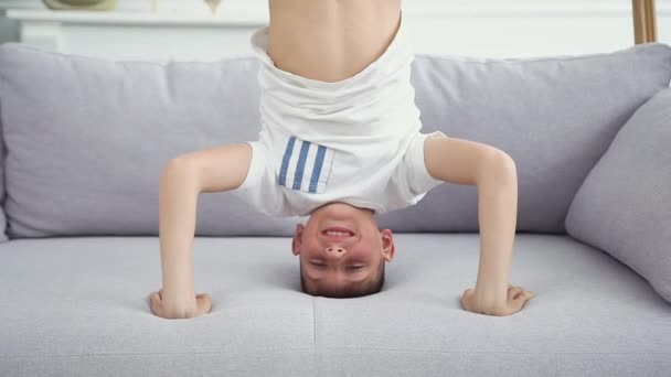 Вид спереду красивий спортивний усміхнений хлопчик-підліток, який стоїть на голові і руках на дивані вдома — стокове відео
