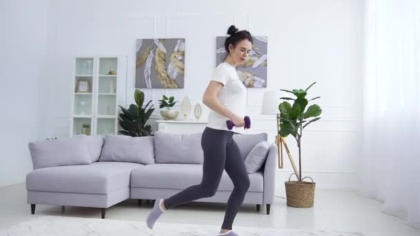 Goed uitziende sportieve zelfverzekerde jonge brunette in sportkleding doet fitness oefeningen met halters thuis — Stockvideo
