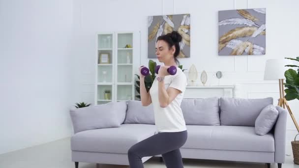 Κοντινό πλάνο του όμορφη θετική νεαρή μελαχρινή σε αθλητικά ρούχα που κάνει ασκήσεις τέντωμα με αλτήρες στο σπίτι — Αρχείο Βίντεο