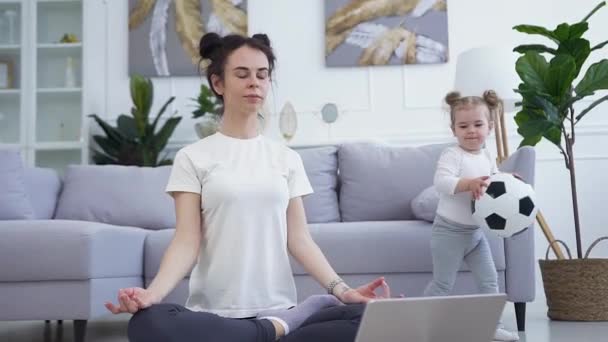 Attraktiv rolig slank ung kvinde sidder på gulvet på lotus udgør mens hendes energiske smukke lille datter leger rundt med bolden – Stock-video