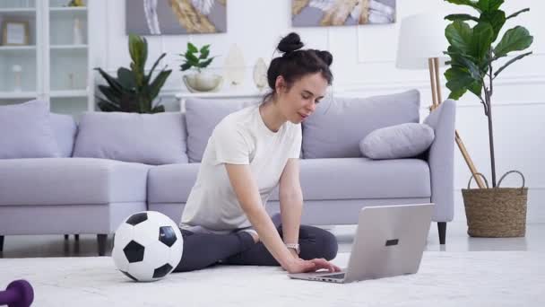 Apelando a la joven delgada satisfecha sentada frente a la computadora portátil y haciendo ejercicios de yoga relajantes en el suelo en la sala de estar — Vídeos de Stock