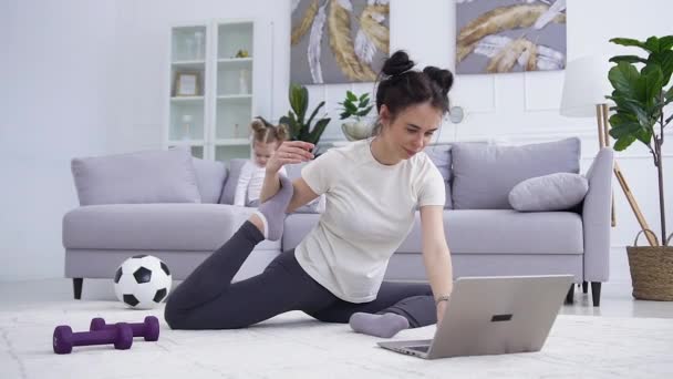 Trevlig nöjd lugn ung kvinna i sportkläder gör stretching yoga övningar på golvet hemma medan hennes söta lilla flicka spelar på soffan — Stockvideo