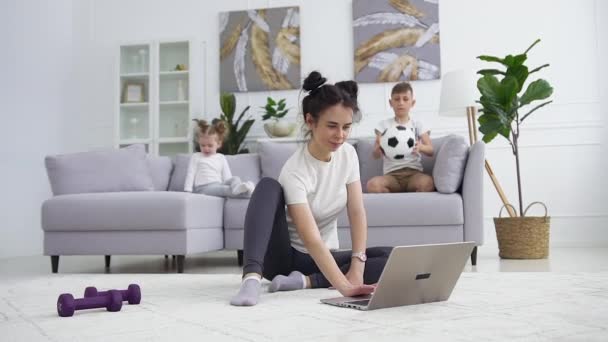 Idílio familiar onde boa aparência calma diferentes idades crianças sentadas no sofá, enquanto sua mãe sorrindo esportivo bonito fazendo exercícios de alongamento de fitness no tapete perto do laptop — Vídeo de Stock