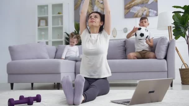 Вид довольной добродушной молодой женщины, которая практикует йогу для растяжки на полу дома, в то время как ее привлекательный сын-подросток и годовалая дочь играют на диване — стоковое видео
