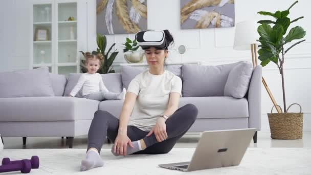 Gadis kecil yang tenang manis duduk di sofa dan menonton ibunya yang duduk di lantai di headset realitas virtual dan berlatih latihan yoga — Stok Video