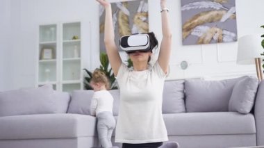 Sanal gerçeklik kulaklığı takmış, yerde yoga egzersizleri yapan sevimli bir yaşındaki kızının yanında oldukça sakin genç bir kadının ön görüntüsü.