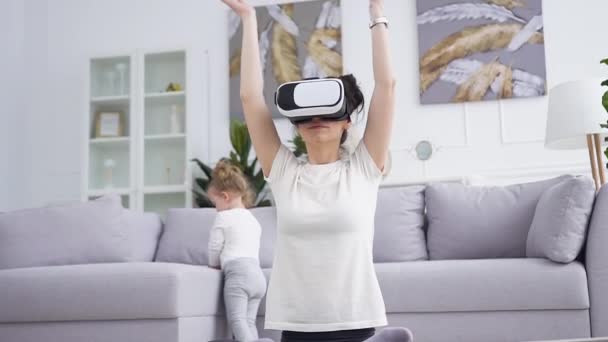 Elülső kilátás elég nyugodt fiatal nő virtuális valóság headset, amely jóga gyakorlatok a padlón mellett az ő aranyos vicces egy éves lánya — Stock videók