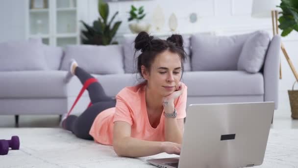 Wanita muda yang bahagia dan damai dengan gaya rambut lucu mengenakan pakaian olahraga melakukan latihan peregangan dengan tali peregangan berbaring di depan laptop di lantai — Stok Video