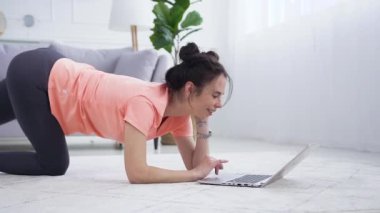 Spor kıyafetli, güzel gülümseyen genç bir kadının yan görüntüsü bilgisayarda spor kompleksi egzersizleri izleyip bacağını evde esnetiyor.