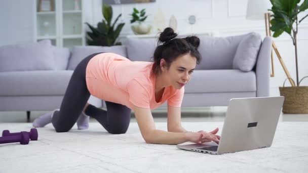 Добродушная довольная молодая женщина в современной прическе наблюдая фитнес-тренировки на ноутбуке и одновременно растягивая ногу — стоковое видео
