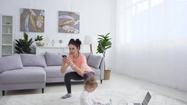 Spor kıyafetleri içinde neşeli, genç bir kadın spor salonunda spor yaparken spor yaparken spor egzersizleri yapıyor. Küçük tatlı kızı da halının üzerinde oynuyor.