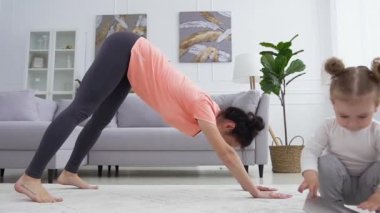 Spor tişörtlü ve taytlı genç bir kadının ön görüntüsü. Evde gülümseyen küçük kızının yanında spor yoga egzersizleri yapıyor.