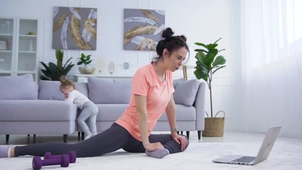 Ελκυστική σπορ νεαρή προπονήτρια γυμναστικής κάθεται στο πάτωμα και εξηγεί τις πολύπλοκες ασκήσεις για την ομάδα γυμναστικής της μέσω βιντεοκλήσης στο laptop — Αρχείο Βίντεο