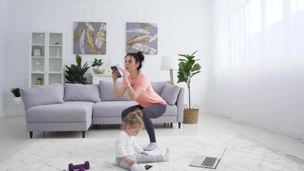 Hermosa niña pequeña sentada en el suelo cerca de la computadora portátil, mientras que su madre esbelta deportiva segura haciendo ejercicios en cuclillas — Vídeos de Stock