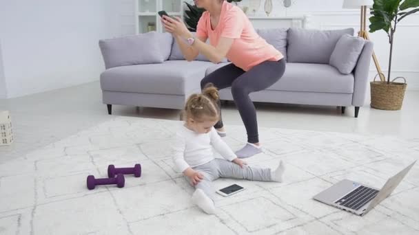 Soustředěná malá dívka sedí na podlaze a sleduje kreslený telefon, zatímco její usmívající se krásná sportovní matka dělá squat cvičení — Stock video