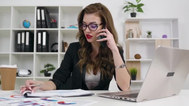 Gros plan d'adorable femme d'affaires moderne souriante qui parle au téléphone tout en travaillant sur un ordinateur portable dans une pièce de bureau magnifiquement conçue, 4k — Video
