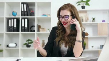 Göz kamaştırıcı mutlu, gülümseyen ofis çalışanı, gözlüklü, parlak makyajlı, ofis odasında telefon konuşmasının keyfini çıkaran.
