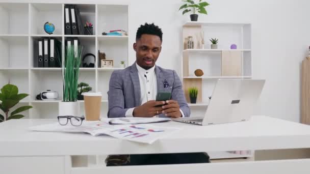 Handsome smiling young african american in business clothes sitting at the table in office and using his smartphone,4k — Stock Video