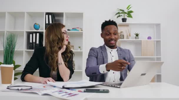 Attraktiv glad leende ung målmedveten ljus kvinna och hennes svarthyade manliga kollega arbetar tillsammans vid kontorsbordet, slow motion — Stockvideo