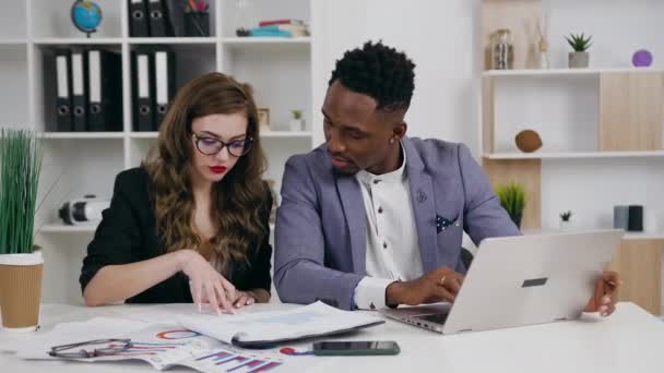 Целеустремленная внимательная опытная молодая предпринимательница и ее чернокожая партнерша, работающая вместе с документами и информацией с ноутбука в офисе, 4k — стоковое видео