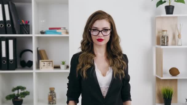 Vista frontal de la impresionante feliz éxito joven y moderna dama de negocios en gafas con estilo de ropa de negocios y con lápiz labial rojo que se encuentra en la habitación de la oficina y posando en la cámara — Vídeo de stock