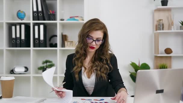 Vorderansicht einer rücksichtsvoll sympathischen, verantwortungsbewussten jungen Geschäftsfrau in Brille in stylischer Kleidung und mit hellem Make-up, die Berichte mit Grafiken im Büroraum überprüft — Stockvideo