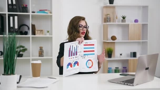 Belle jeune femme d'affaires souriante et satisfaite montrant un projet d'affaires à son interlocuteur par vidéoconférence dans un bureau contemporain — Video