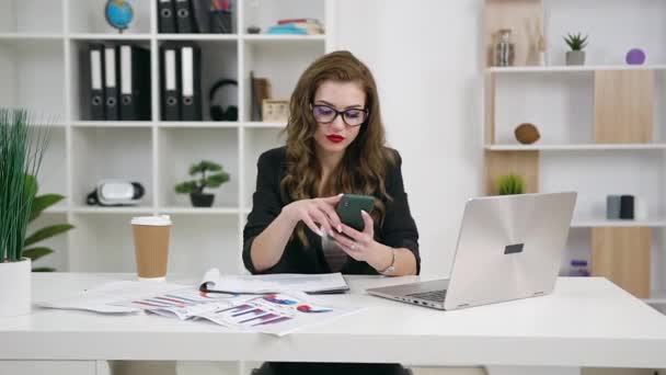 Γοητευτική αυτοπεποίθηση συγκεντρωμένη νεαρή γυναίκα διευθυντής γραφείου σε γυαλιά, ντυμένος με κομψά ρούχα κάθεται στο χώρο εργασίας της και χρησιμοποιώντας smartphone — Αρχείο Βίντεο