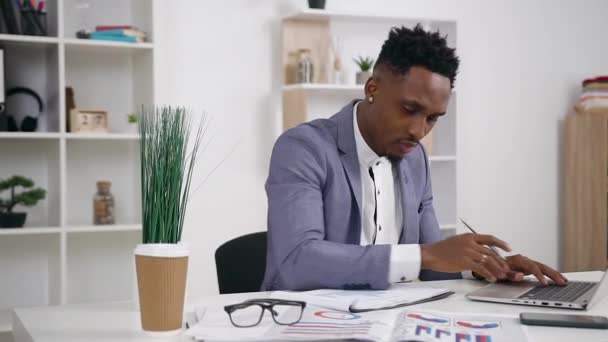 Handsome purposeful dark-skinned young male office worker typing on laptop and writing down to his report important marks — Stock Video