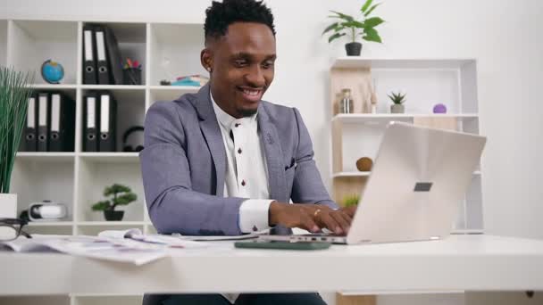 Attraktives Lächeln zufriedene hochqualifizierte dunkelhäutige Büroleiter sitzt an seinem Arbeitsplatz und arbeitet am Laptop, Nahaufnahme — Stockvideo
