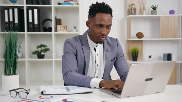 Handsome positive smiling young black-skinned office manager posing on camera with sincerely smile while working on laptop in office — Stock Video