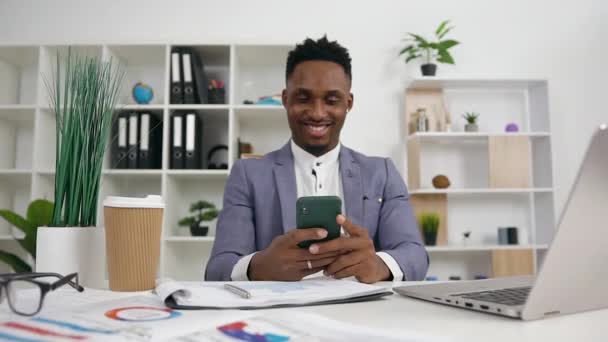 Elülső kilátás vidám jóképű nyugodt, fiatal, fekete bőrű férfi irodavezető stílusos viselet, amely használja a telefont ül a munkahelyén az irodában — Stock videók