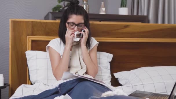 Perturbada jovem morena sentada na cama do quarto com cachecol ao redor do pescoço falando ao telefone enquanto trabalhava remotamente durante a doença da gripe — Vídeo de Stock