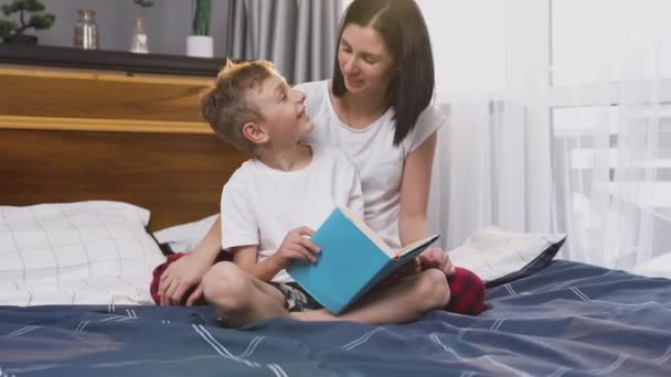 Вид на привлекательную довольную любящую женщину и ее 8-летнего сына, которые веселятся вместе, читая книгу в спальне — стоковое видео