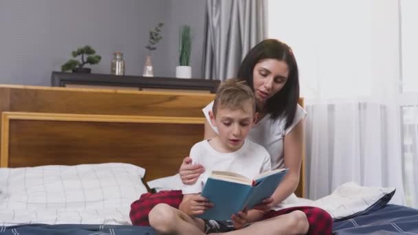 Concepto familiar donde el guapo niño sonriente leyendo el libro junto con su hermosa madre alegre de pelo oscuro en el dormitorio — Vídeos de Stock