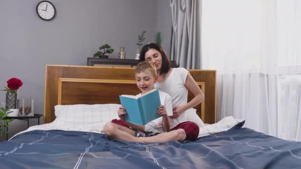 Atractivo feliz niño de 8 años leyendo libro en la cama del dormitorio mientras su madre sonriente y cariñosa le cosquillas — Vídeo de stock