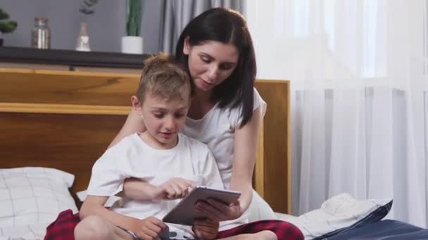 Schließen Sie die gut aussehende lächelnde glückliche Familie von Mutter und Sohn, die zu Hause auf einem bequemen Bett sitzt und Tablet-PC benutzt — Stockvideo